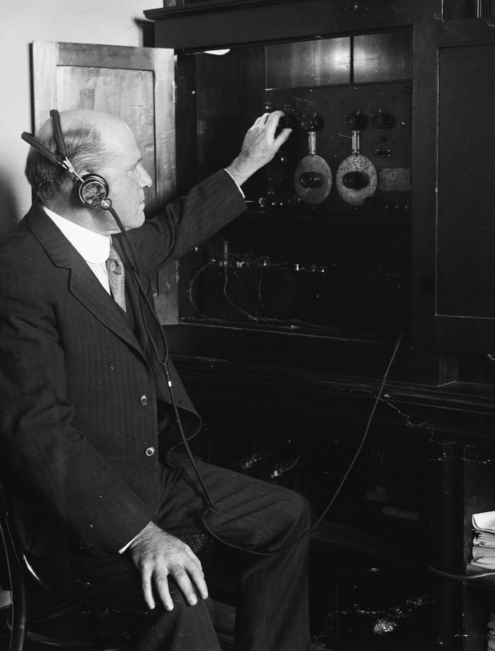A black and white photograph of a man in a suit and headphones, seated, reaching up to tune the dials of a large cabinet wireless set